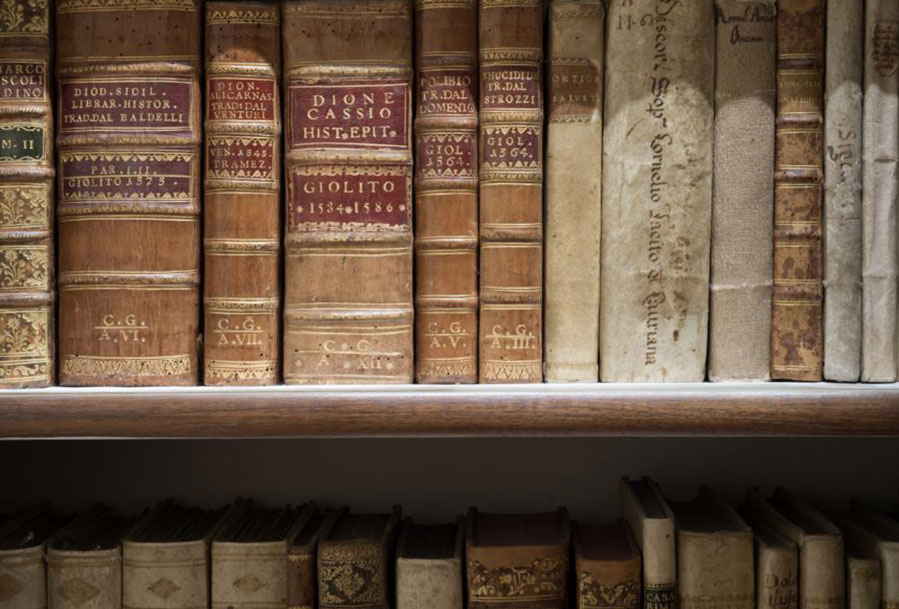 L'immagine mostra uno scaffale con libri antichi rilegati in cuoio, etichettati con titoli storici e classici, indicandone l'antichità.