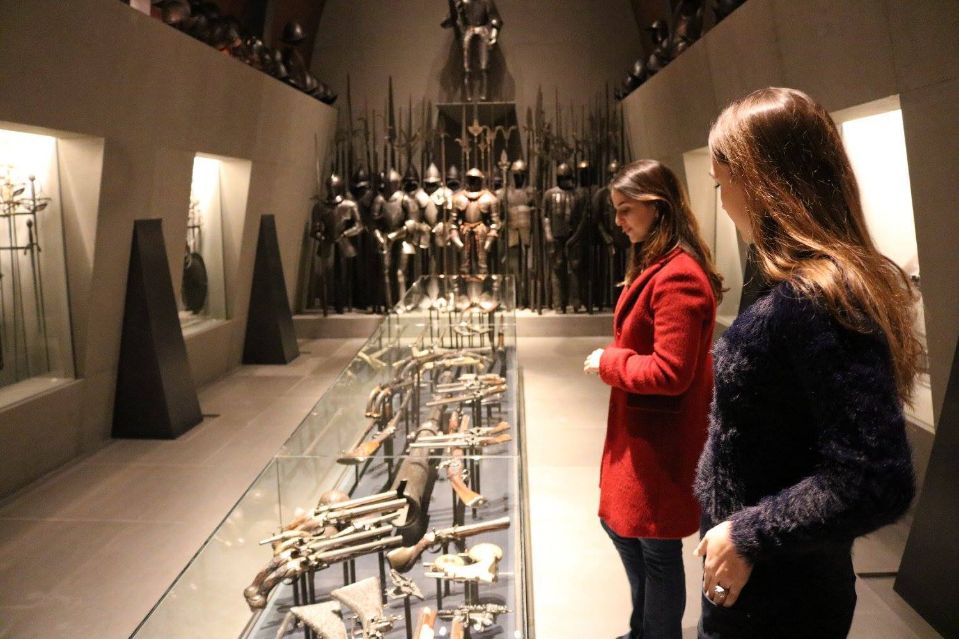 Due persone osservano esposizioni nella Sala d'Armi del Museo Poldi Pezzoli, armature e armi in mostra dentro vetrine e sulla parete del fondo.
