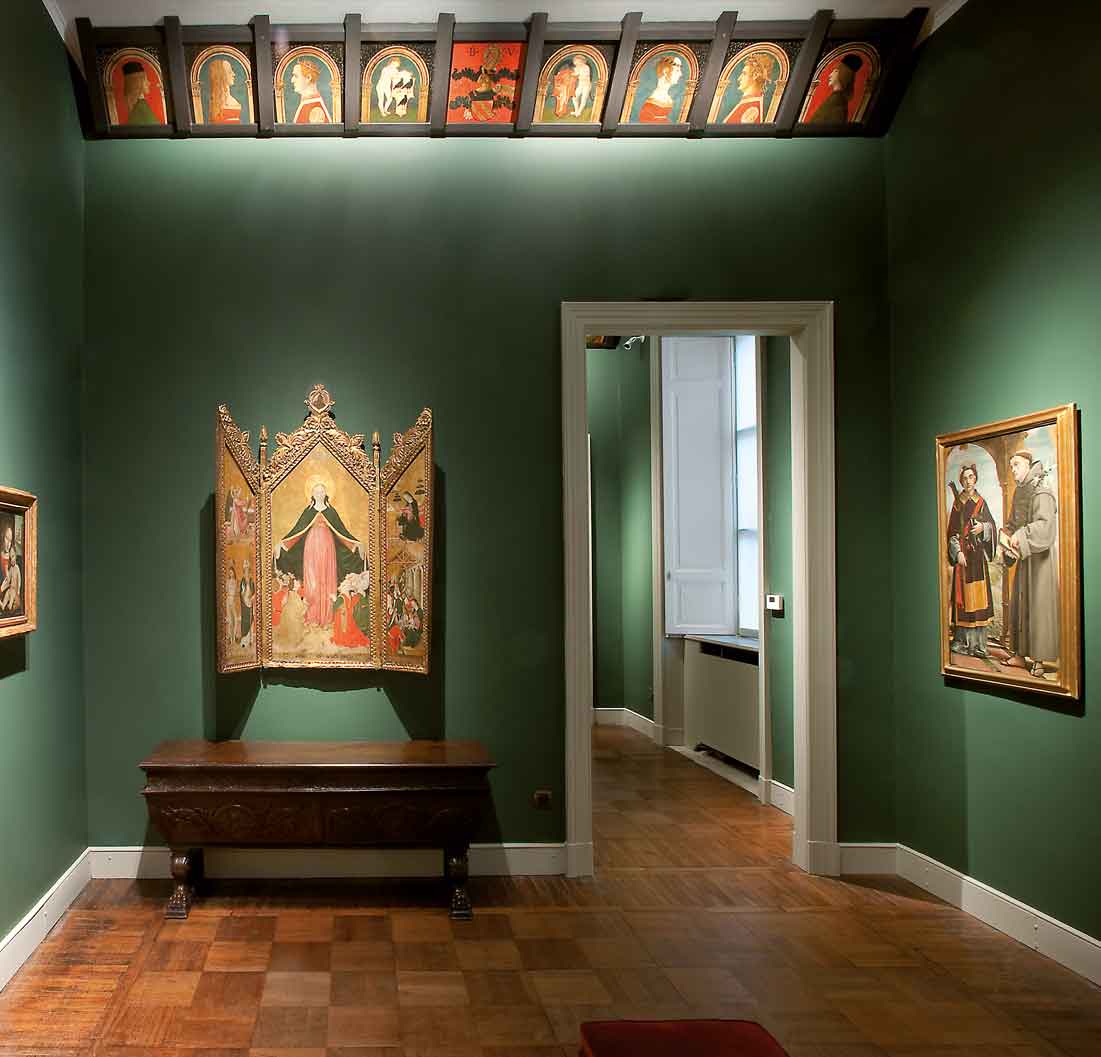 Interno di un museo con pareti verdi e opere d'arte. Un dipinto religioso centrale su leggio, cornici con ritratti laterali e decorazioni medievali in alto.