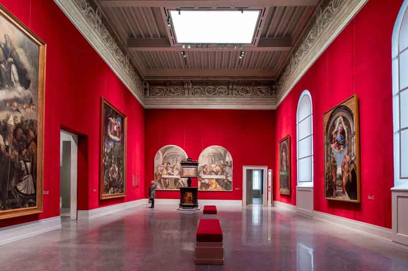 Sala della Pinacoteca Tosio Martinengo di Brescia con pareti rosse, quadri di grandi dimensioni, soffitto alto, pavimento in legno e una persona che osserva.