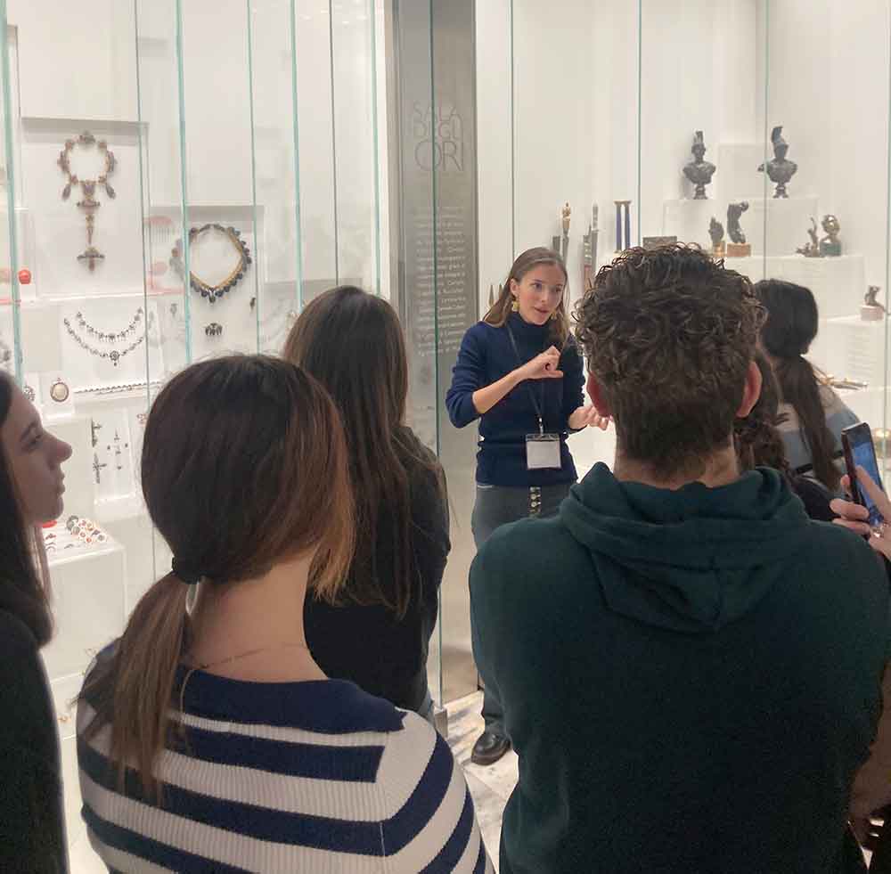 Un gruppo di persone ascolta una guida al museo Poldi Pezzoli mentre indica gioielli esposti in vetrine. Ambiente interno, illuminazione soffusa, atmosfera educativa.