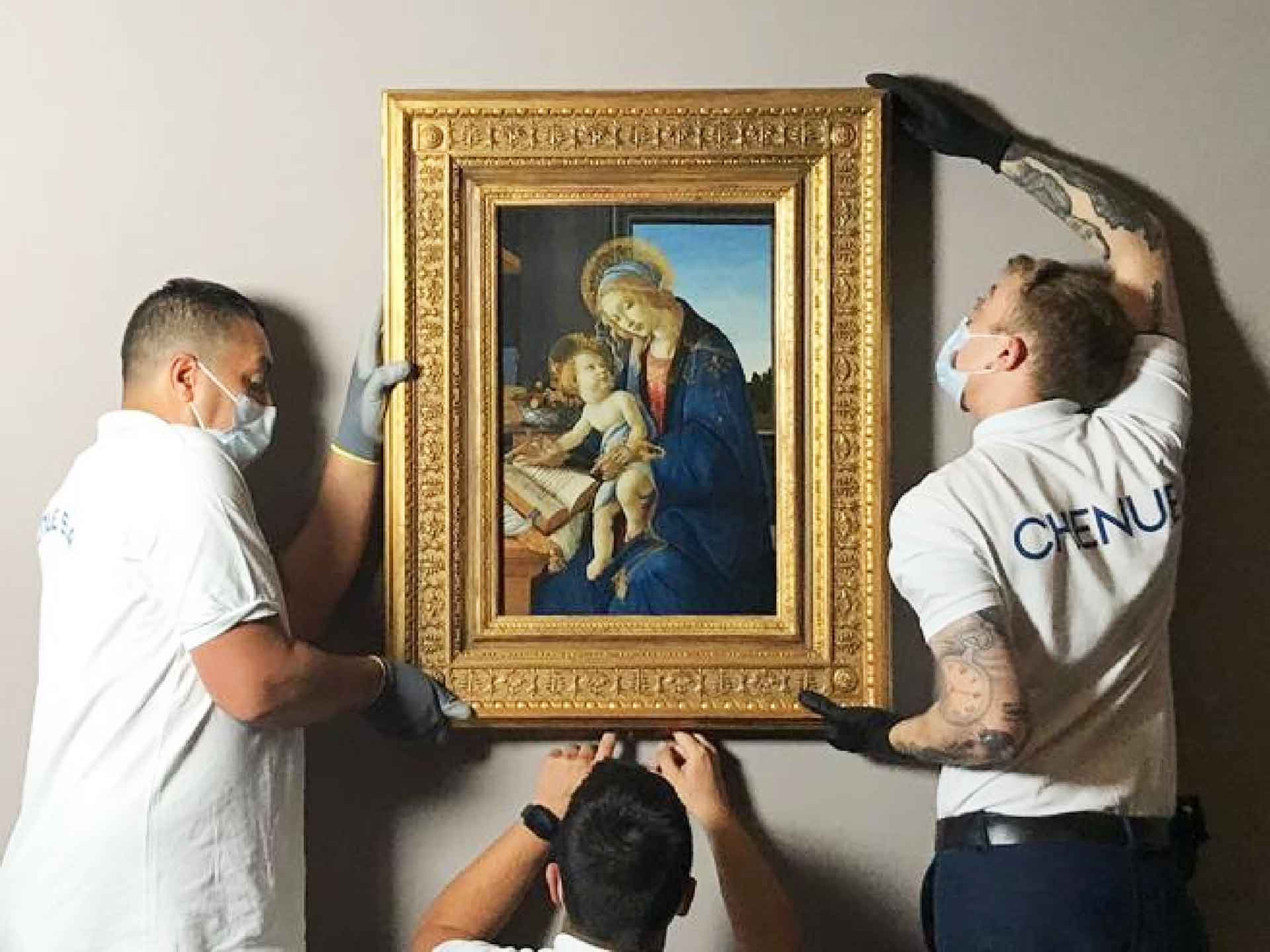 Tre professionisti stanno appendendo un quadro di Botticelli raffigurante la Madonna con Bambino. Sono attenti e indossano guanti per proteggere l'opera d'arte.