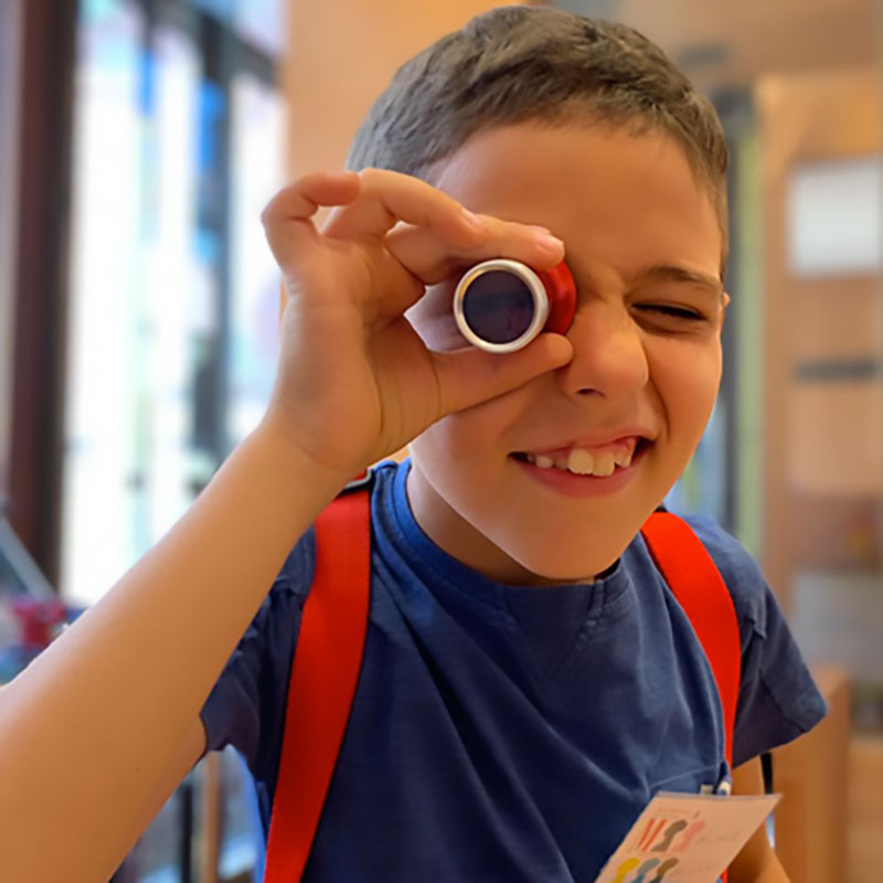Un bambino sorridente guarda attraverso un piccolo oggetto cilindrico rosso, indossa una maglietta blu e uno zaino con spallacci rossi.