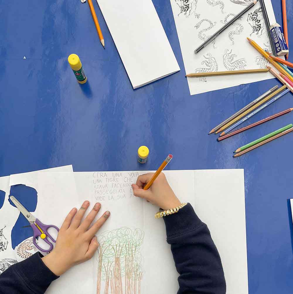 Un bambino sta disegnando con matite colorate su un foglio di carta. Sul tavolo ci sono forbici, matite, e colla. 