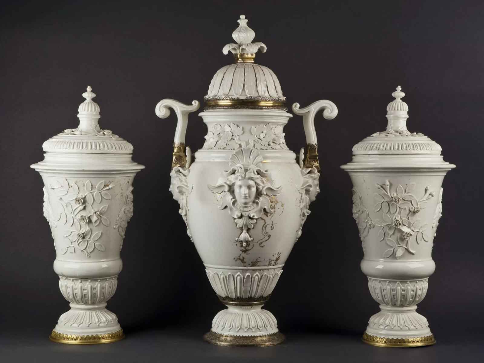 Tre grandi vasi in porcellana bianca con dettagli decorativi rilievi floreali e volti, manici eleganti e accentuazioni dorate su base scura.
