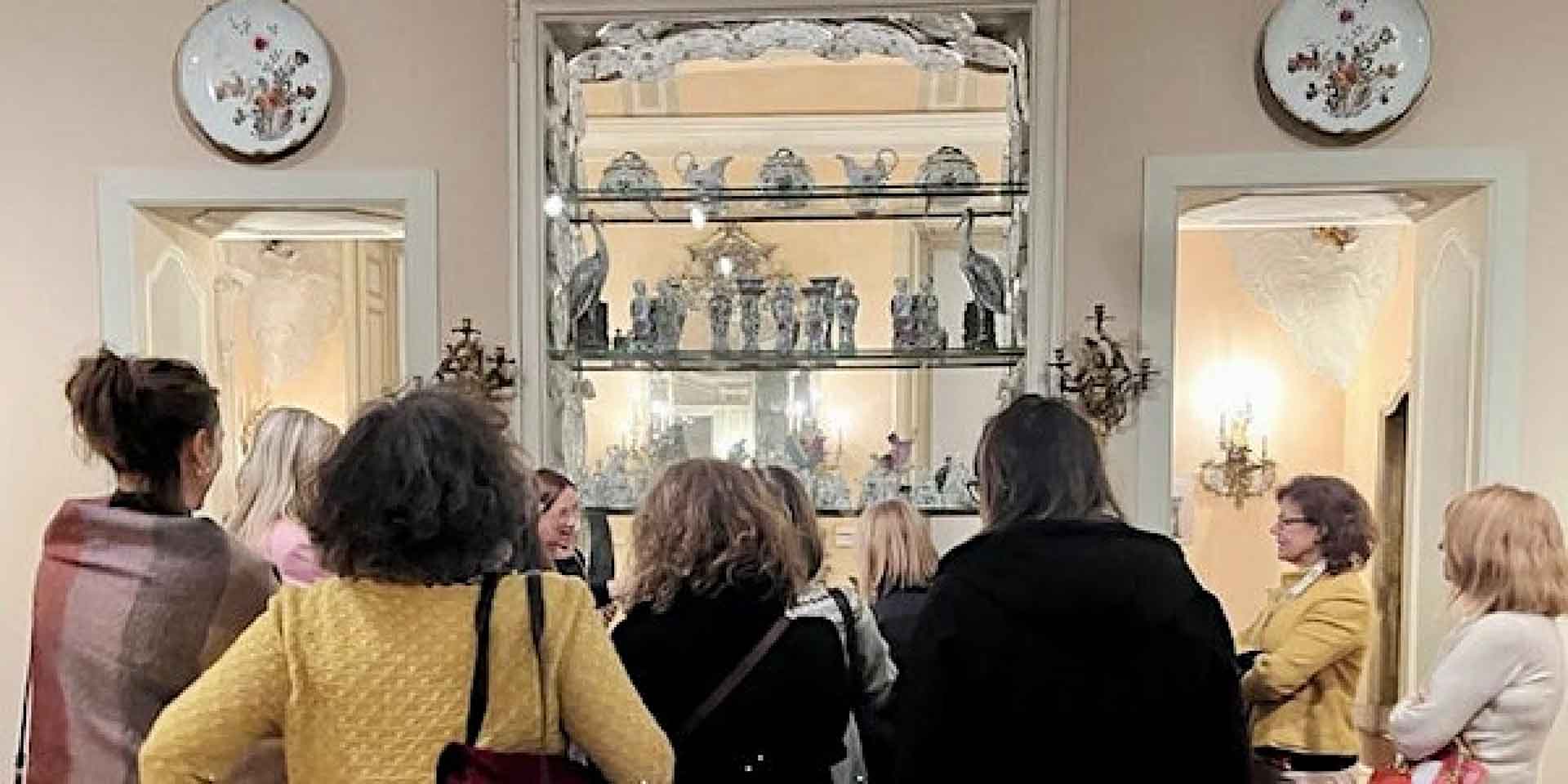 Un gruppo di persone osserva interessato degli oggetti esposti in una vetrina all'interno di una stanza decorata con specchi e piatti ornamentali.