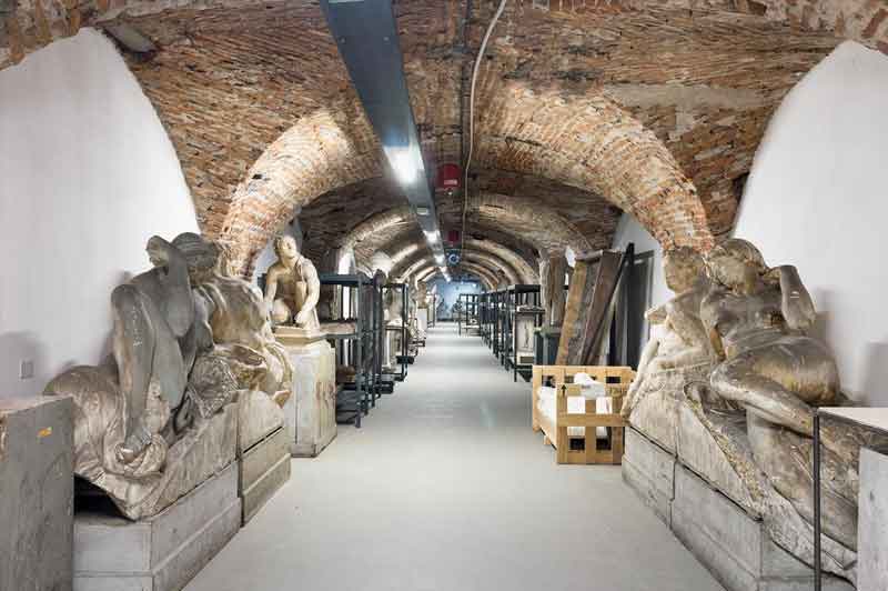 Accademia di Brera a Milano Un corridoio con soffitto a volta in mattoni, sculture posizionate lateralmente e quadri impilati. L'atmosfera è quella di un deposito o archivio d'arte.