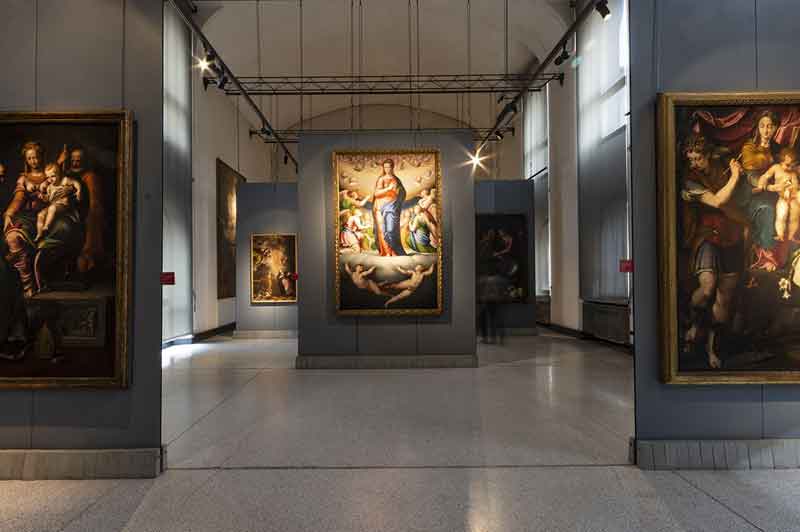 Il Museo Ala Ponzone a Cremona con pareti grigie, pavimento lucido e quadri incorniciati appesi, illuminati da faretti a soffitto. Nessuna persona visibile.