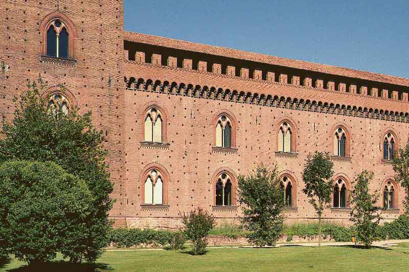 Edificio dei Musei Civici di Pavia  con mattoni a vista, finestre ad arco, merlatura lungo il tetto e circondato da alberi verdi su erba ben curata.