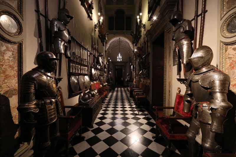Un corridoio del Museo Bagatti Valsecchi di Milano con pavimento a scacchiera, armature medievali in fila lungo le pareti ornate e busti su piedistalli, atmosfera storica e illuminazione soffusa.