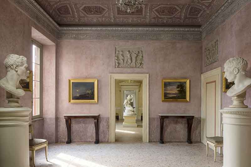 Elegante stanza interna di Palazzo Tosio Brescia con pareti rosa, soffitto decorato, sculture busti, dipinti in cornici dorate, specchi, e una serie di porte aperte.