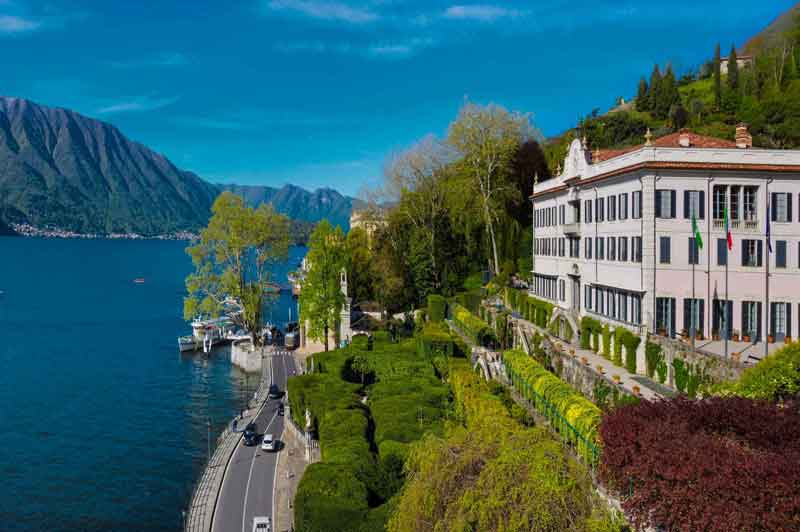 Villa Carlotta elegante sul lago con giardini terrazzati, montagne sullo sfondo, cielo azzurro e alcune auto sulla strada adiacente.